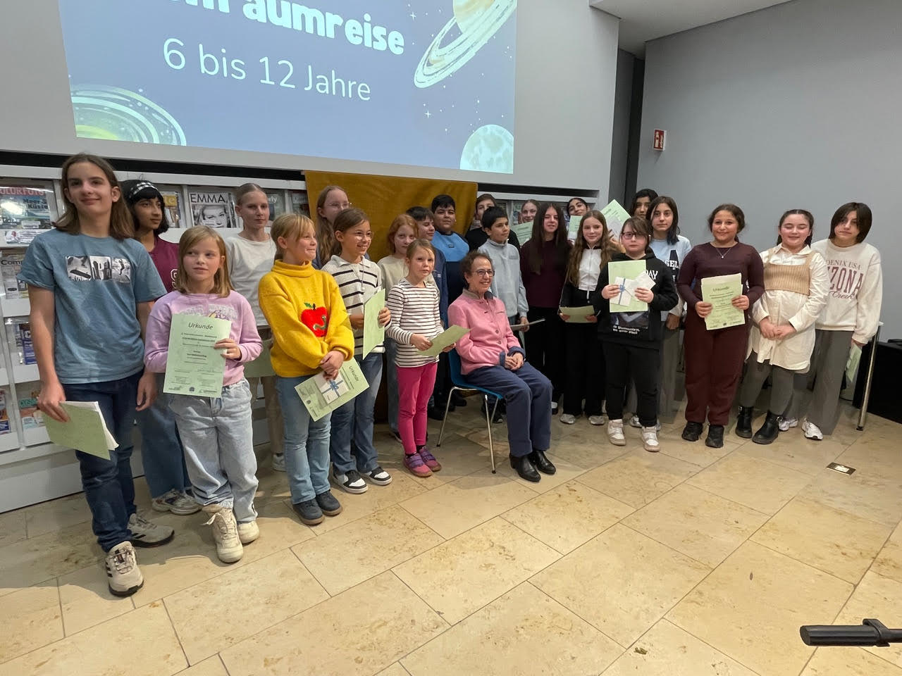 Jugendpreis 2025 6 bis 12, Auf der linken Seite die Teilnehmerinnen und Teilnehmer am Wettbewerb „Weltraumreise“ in der Mitte sitzt die Stifterin der Arno Reinfrank  Jugendpreises Frau Jeanette Koch -Reinfrank und auf der rechten Seite sehen wir die anwesenden Teilnehmerinnen und Teilnehmer aus der Kategorie Bildergeschichte ( F&ouml;rderklasse im Fach Deutsch)